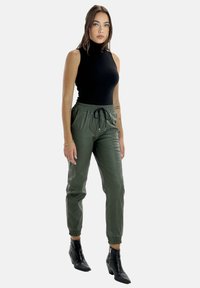 Schwarzes ärmelloses Top, grüne Jogginghosen mit elastischem Bund und schwarzen Stiefeletten. Glattes Material mit schmal zulaufenden Beinen und Kordelzugdetails.