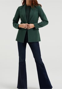 Grön skräddarsydd blazer med slagkrage, enda knappöppning och två framfickor, tillsammans med utsvängda mörk denimjeans.