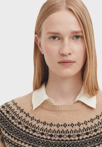 Strickpullover in hellem Beige mit einem schwarzen gemusterten Einsatz, runder Ausschnitt, über einem weißen Hemd mit Kragen getragen. Glatte Textur, lässiges Design.