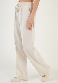Beige linnen lange broek met een elastische tailleband en koord, met zijplooien en een relaxte pasvorm. Gepaard met lichtgekleurde sneakers.