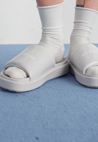 Grå slidesandaler med en bred stropp som har teksten "AIR JORDAN", brukt over hvite sokker, plassert mot en blå teppelbunn.