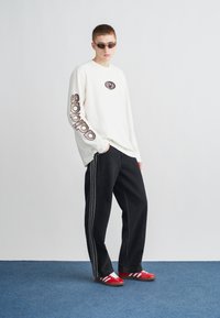 Hosszú ujjú krémszínű póló nyomtatott logóval, fekete farmerrel, oldalcsíkkal, és piros csíkos tornacipővel párosítva. Sötétített lencsés szemüveg.