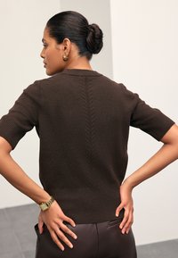 Femme aux cheveux foncés en chignon porte un pull marron à manches courtes et une montre en or, debout, les mains sur les hanches, dos à la caméra.