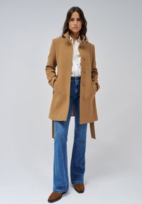 Light brown wool blend coat with a tailored fit, 5 buttons, side pockets, worn over a white blouse and blue flared jeans; brown boots.