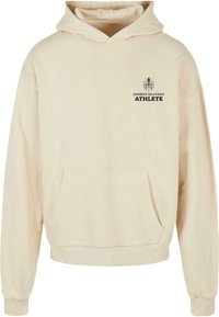 Beige hoodie gemaakt van zachte stof, met een voorzak en een capuchon met trekkoord. Zwarte print met het logo "SPORTS SEASON ATHLETE" op de borst.