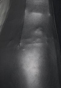 Material de couro preto com uma textura suave, apresentando um detalhe de costura ao longo do lado e um leve brilho que reflete a luz.