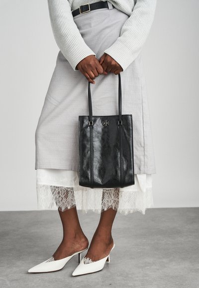 Borsa tote in pelle nera con due manici, texture liscia e dettaglio con logo. Abbinata a una gonna grigia chiara con orlo in pizzo bianco.