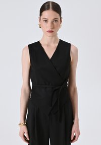 Top nero avvolgente in tessuto morbido, caratterizzato da un profondo scollo a V, design senza maniche e vita annodata. Abbinato a orecchini geometrici dorati.