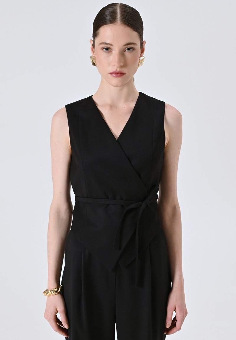 Top nero avvolgente in tessuto morbido, caratterizzato da un profondo scollo a V, design senza maniche e vita annodata. Abbinato a orecchini geometrici dorati.