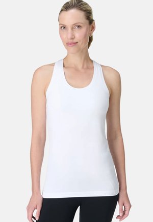 Vrouw met naar achteren gebonden haar, gekleed in een witte, mouwloze sporttop en zwarte legging, staand tegen een effen lichte achtergrond.