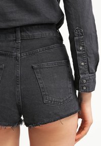 Shorts de mezclilla negros con dobladillo deshilachado y bolsillos traseros, combinados con una camisa de manga larga a juego que presenta detalles de botones en las mangas.