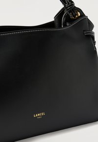 Μαύρη δερμάτινη τσάντα χειρός με λευκή ραφή, χρυσαφένιες λεπτομέρειες και το λογότυπο "LANCEL PARIS" ανάγλυφο σε χρυσό στο μπροστινό μέρος.