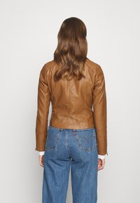 Brun læderjakke med tætsiddende design, der har skuldersyninger, lange ærmer og lynlåse ved ærmekanterne. Båret over blå jeans.