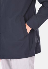 Giacca impermeabile blu navy con una texture liscia, tasche laterali e una vestibilità leggermente rilassata. Presenta un alto colletto e polsini elastici.