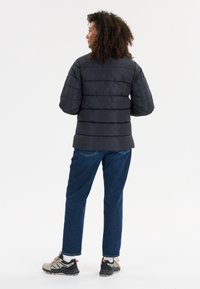 Svart vadderad pufferjacka med horisontella ränder, tillsammans med blå denimbyxor och gråsvarta sneakers. Bakgrunden är helt vit.