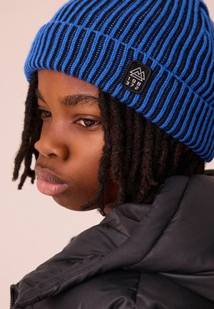Bonnet en tricot rayé bleu et noir avec un petit logo de montagne noir. La personne a des cheveux sombres et bouclés et porte une veste noire rembourrée.