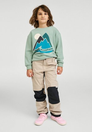 Jasnozielona bluza z nadrukiem gór, w połączeniu z beżowymi spodniami z czarnymi akcentami. Dziecko ma na sobie różowe trampki.