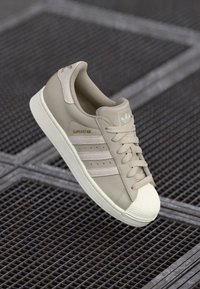Os ténis bejes Adidas Superstar apresentam três riscas horizontais, um design em pele texturizada, biqueira em borracha e marcação dourada lateral.
