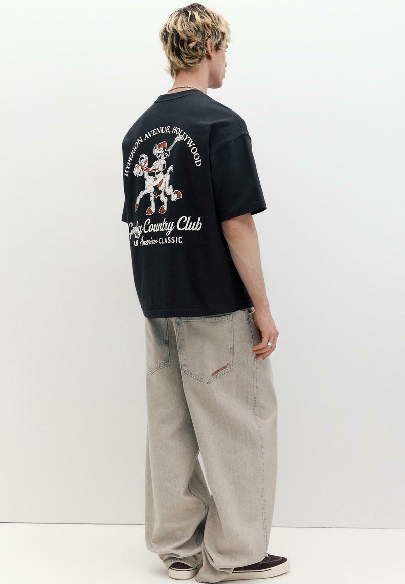 Jeune homme aux cheveux blonds portant un t-shirt noir à motifs graphiques et un jean large gris clair, debout contre un fond blanc, de profil.