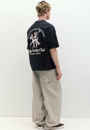 Junger Mann mit blondem Haar, trägt ein schwarzes T-Shirt mit Grafik und weite, hellgraue Jeans, steht vor weißem Hintergrund und blickt seitlich.