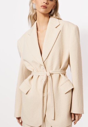 Frau trägt texturierten cremefarbenen Blazer mit Gürtel, der in der Taille gebunden ist, und weit ausgeschnittenen Revers, kombiniert mit großen quadratischen Strassohrringen.