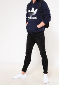 Námořnická modrá mikina s bílým logem Adidas, černé slim-fit džíny a bílé tenisky. Oblečeno s černým batohem. Neformální outfit.