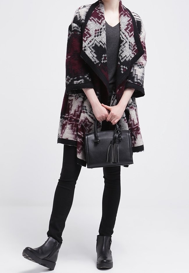 Personne portant un cardigan ouvert à motifs bordeaux, noir et blanc sur un haut gris, un pantalon noir, des bottines noires, tenant un sac à main noir avec des pampilles.