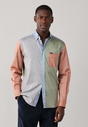 Man wearing a long-sleeve button-up shirt with color blocks in blue, gray, green, and orange, posing with one hand in pocket.