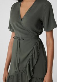 Robe portefeuille vert kaki en tissu lisse, avec de courtes manches volantes, une accentuation froncée et un détail de taille nouée.