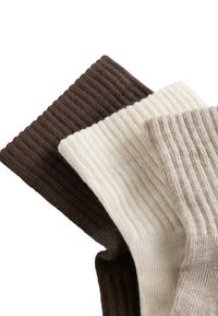 Három összehajtott bordázott kötött zokni sötétbarna, krémszínű és világos bézs árnyalatokban, egymásra halmozva fehér háttéren.