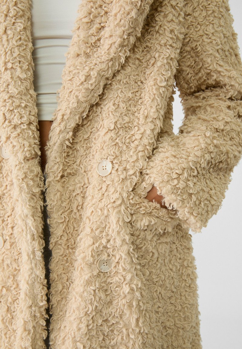 Beżowy puszysty płaszcz z teksturowanym, kręconym materiałem. Posiada szalowy kołnierz, dwa guziki i boczne kieszenie.