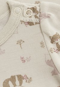 Ett babytäcke i bomull med en krämfärgad bakgrund som har små illustrationer av tvättbjörnar och blommor i beige och ljusrosa. Tryckknappar.