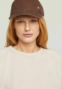 Brown cap with a curved brim, cotton material, logo detail on the front, paired with a light beige sweatshirt featuring a ribbed neckline.