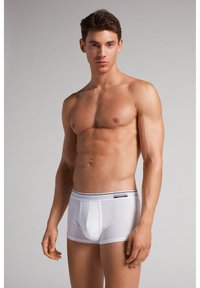 Jonge gespierde man staand, gekleed in strakke witte boxer briefs met merkband, kijkend naar voren tegen een effen neutrale achtergrond.