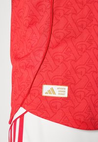 Crvena sportska majica s teksturiranom tkaninom koja ima ponavljajući uzorak logotipa, ukrašena zlatnom etiketom s natpisom "AUTENTIČNI DOZVOLJENI PROIZVOD."
