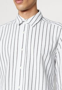 Witte overhemdresshirt met verticale grijze strepen, kraag en knoopsluiting aan de voorkant. De stof lijkt lichtgewicht en gestructureerd.