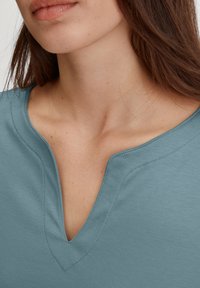 Lichtblauwe V-hals top van zachte stof, met gestikte details langs de halslijn en een gladde, licht getextureerde oppervlakte.