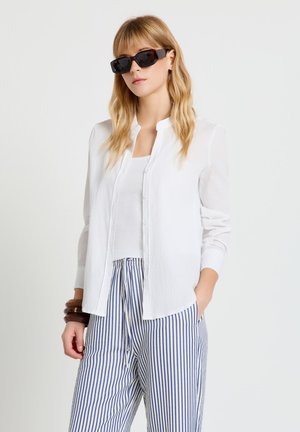 Donna con occhiali da sole neri, camicia bianca abbottonata sopra una maglietta bianca e pantaloni a righe verticali blu e bianche, in piedi con una mano in tasca.