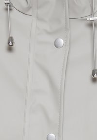 Gros plan sur une veste gris clair avec des boutons-pression argentés et des cordons blancs aux embouts métalliques argentés.