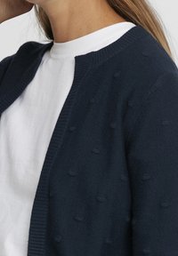 Marinblå stickad kofta med ett strukturerat mönster och en ribbad krage. Bärs över en vit t-shirt med rund halsringning, vilket visar en lagerstil.