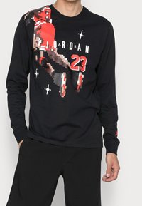 Mies, joka pukeutuu mustaan pitkähihaiseen paitaan, jossa on pikselöity koripalloilijan grafiikka, teksti "JORDAN" ja numero 23 punaisella ja valkoisella.