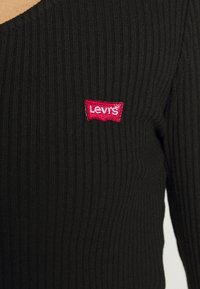 Černý žebrovaný top s designem ve tvaru V a červeným vyšívaným logem Levi's na hrudi. Tkanina vykazuje jemnou vertikální texturu.