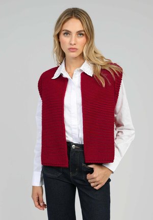 Femme aux cheveux blonds portant une chemise blanche boutonnée, un gilet sans manches en grosse maille rouge et un jean foncé, debout devant un fond gris.
