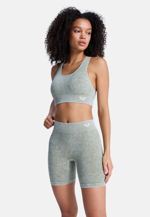 Vrouw draagt een lichtgroene geribbelde sportbeha en biker shorts die bij elkaar passen, staat met één hand op de heup en kijkt naar de zijkant.