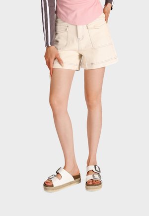 Gambe con pantaloncini color crema con grandi tasche, un top rosa e sandali platform bianchi con fibbie su sfondo chiaro.