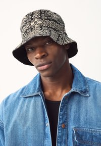 Cappello a secchiello nero con motivi paisley bianchi, realizzato in tessuto; indossato con una giacca di jeans con chiusura a bottoni e tasca sul petto.