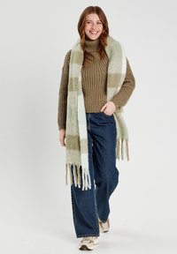 Strikket sweater i olivengrøn, parret med et stort, mønstret tørklæde i lys grøn og hvid. Bredbenede jeans og afslappede sneakers fuldender looket.