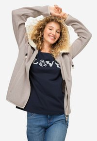 Chaqueta de cremallera gris claro con forro color crema, con mangas largas y un corte relajado, llevada sobre una camiseta azul marino con un diseño estampado.
