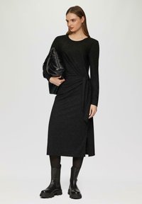 Schwarzes Langarmkleid aus strukturiertem Stoff, tailliert mit gebundenem Detail und einem seitlichen Schlitz; kombiniert mit einer schwarzen Umhängetasche und klobigen schwarzen Stiefeln.