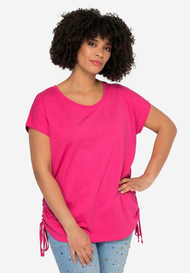 Camiseta corta de manga corta en un vibrante color rosa con escote redondeado y cordones laterales. Tejido suave con un corte relajado. Combinada con unos vaqueros claros bordados.